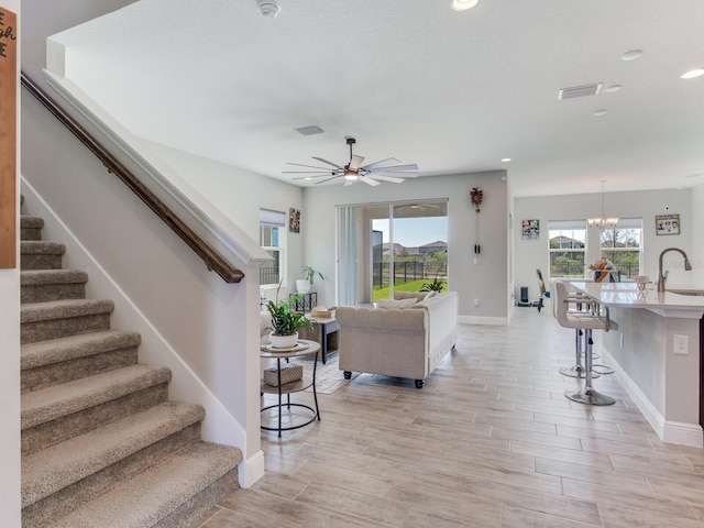 living room featuring ceiling fan with notable chandelier, visible vents, baseboards, stairs, and light wood-type flooring