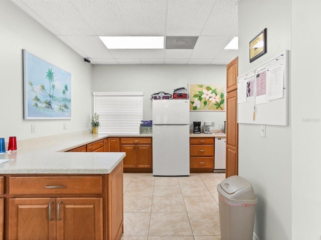 kitchen with light tile patterned floors, a drop ceiling, white appliances, light countertops, and brown cabinetry