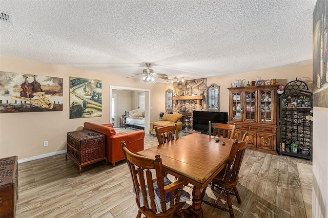 dining room featuring ceiling fan, a stone fireplace, a textured ceiling, and light wood-type flooring