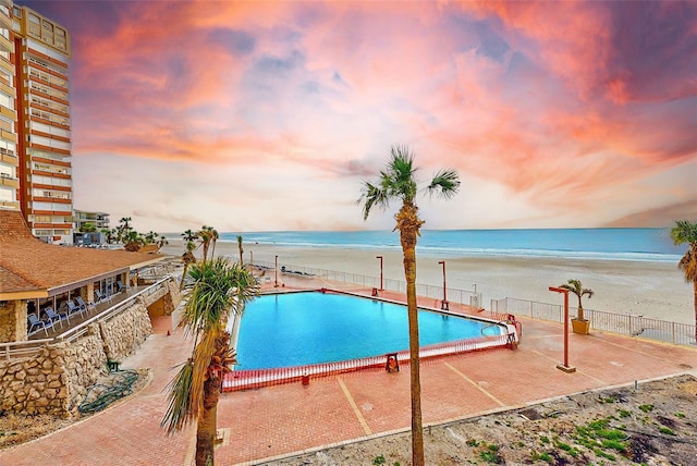 pool at dusk featuring a patio, a community pool, a beach view, a water view, and fence