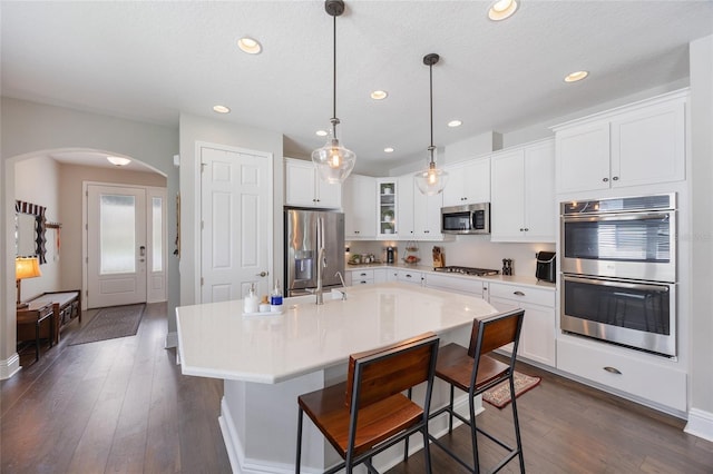 kitchen with appliances with stainless steel finishes, arched walkways, dark wood-style flooring, and glass insert cabinets