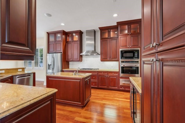 kitchen with a center island with sink, backsplash, light wood-style flooring, appliances with stainless steel finishes, and wall chimney exhaust hood