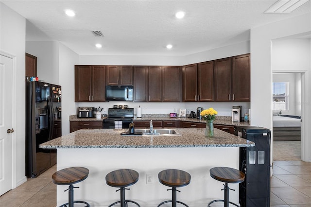 kitchen with dark brown cabinetry, visible vents, light stone countertops, black appliances, and a sink