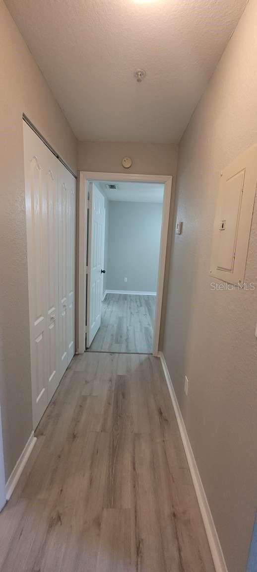 corridor featuring a textured ceiling, light wood-type flooring, electric panel, and baseboards