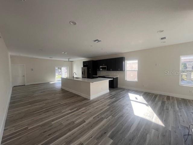 kitchen featuring wood finished floors, open floor plan, dark cabinetry, stainless steel microwave, and an island with sink