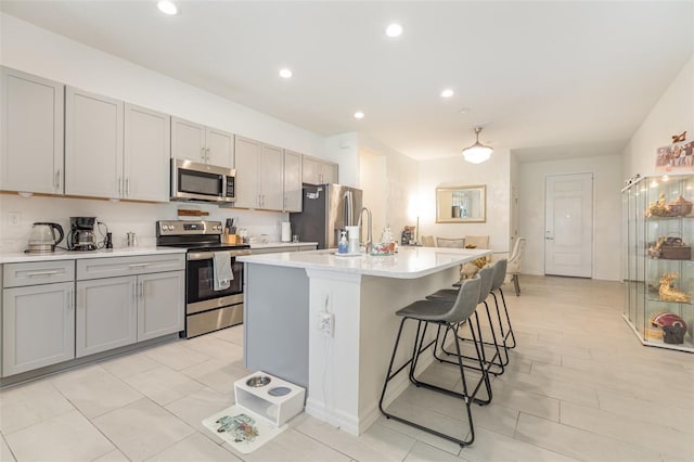 kitchen featuring a center island with sink, appliances with stainless steel finishes, light countertops, gray cabinetry, and a kitchen bar