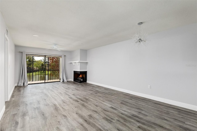 unfurnished living room featuring ceiling fan with notable chandelier, a lit fireplace, wood finished floors, and baseboards