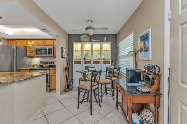 kitchen featuring light tile patterned floors, ceiling fan, stone counters, appliances with stainless steel finishes, and brown cabinets