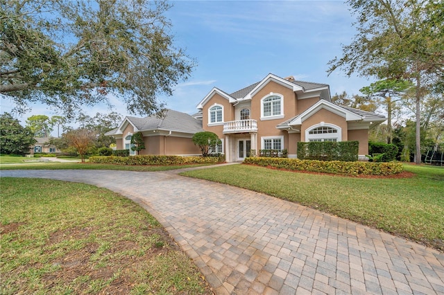 traditional-style home with a front yard, decorative driveway, a balcony, and stucco siding