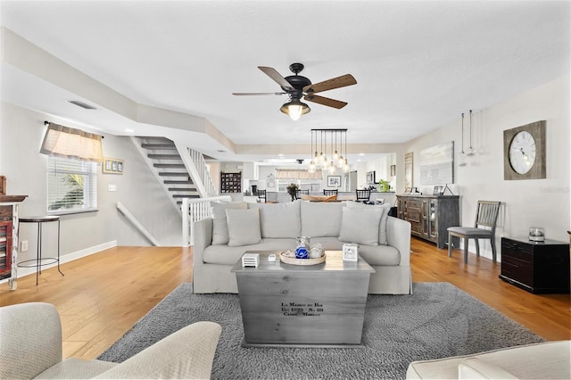 living area with visible vents, baseboards, light wood-style flooring, stairway, and ceiling fan with notable chandelier