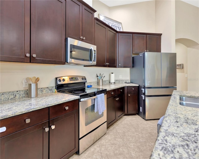 kitchen featuring stainless steel appliances, arched walkways, dark brown cabinetry, light tile patterned flooring, and light stone countertops
