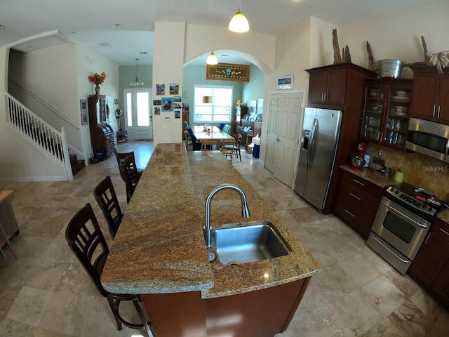 kitchen featuring light stone counters, a sink, appliances with stainless steel finishes, a center island with sink, and glass insert cabinets