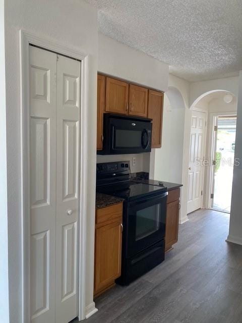 kitchen featuring arched walkways, dark wood-style flooring, dark countertops, brown cabinetry, and black appliances