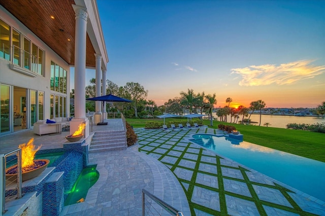 patio terrace at dusk featuring an outdoor fire pit, an outdoor pool, a water view, and a lawn