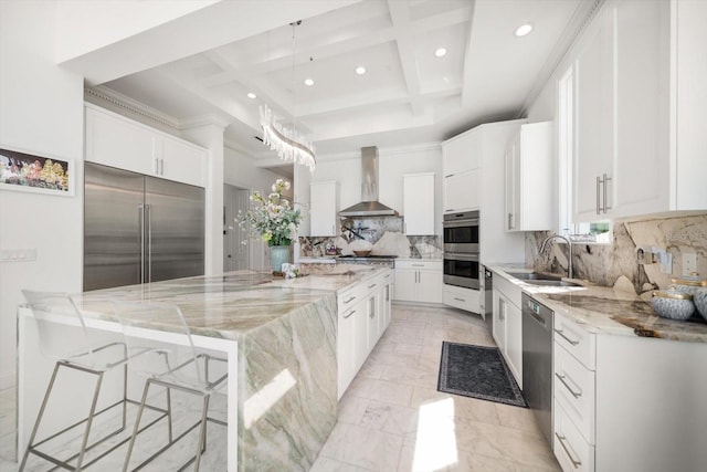 kitchen featuring light stone counters, coffered ceiling, a sink, appliances with stainless steel finishes, and wall chimney exhaust hood