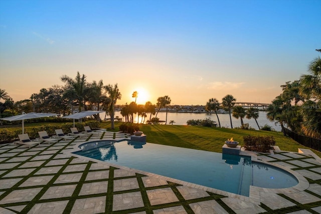 pool at dusk featuring a yard, a patio, a water view, and an outdoor pool