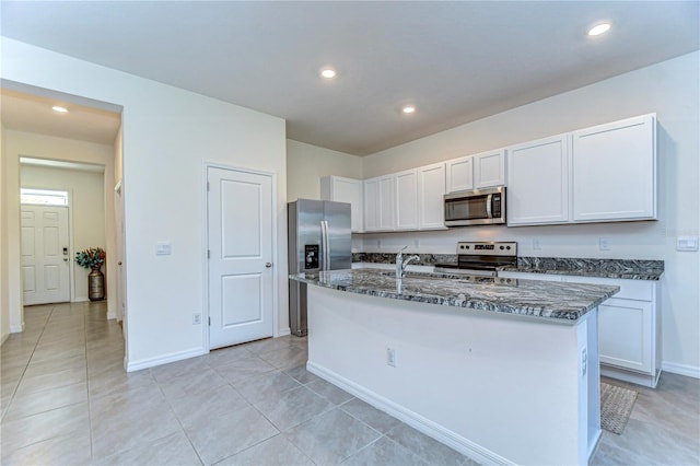 kitchen featuring light tile patterned floors, dark stone counters, an island with sink, appliances with stainless steel finishes, and white cabinetry