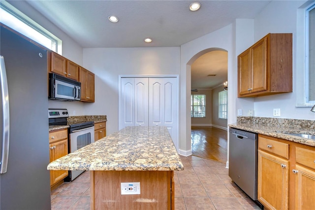 kitchen with light tile patterned floors, arched walkways, a center island, light stone countertops, and stainless steel appliances