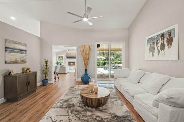 living area with baseboards, ceiling fan, wood finished floors, a lit fireplace, and vaulted ceiling