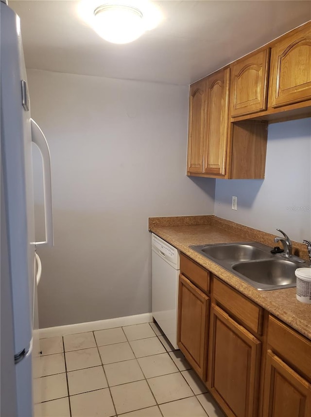 kitchen with light tile patterned floors, brown cabinetry, a sink, white appliances, and baseboards