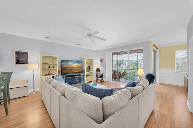 living area featuring ceiling fan, a wainscoted wall, ornamental molding, light wood finished floors, and a glass covered fireplace