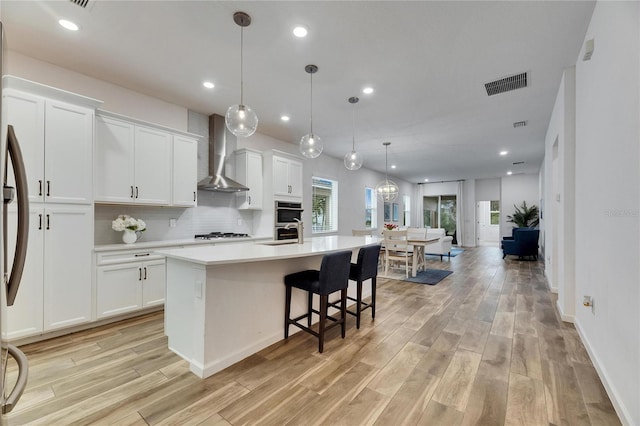 kitchen with an island with sink, wall chimney range hood, visible vents, and light countertops