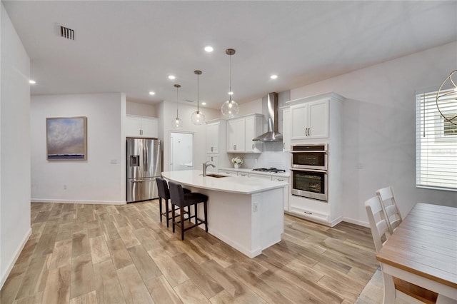 kitchen with light wood finished floors, tasteful backsplash, appliances with stainless steel finishes, a kitchen island with sink, and wall chimney exhaust hood