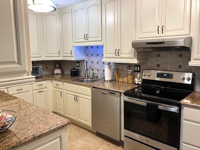 kitchen featuring under cabinet range hood, stainless steel appliances, a sink, backsplash, and light stone countertops