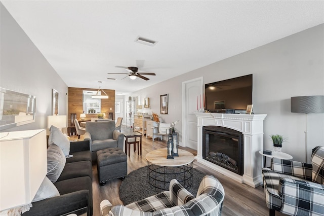 living area featuring visible vents, a glass covered fireplace, ceiling fan, wood walls, and wood finished floors