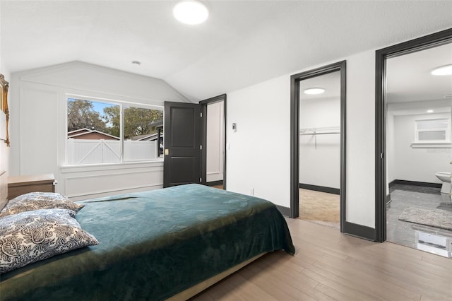 bedroom with a walk in closet, light wood finished floors, ensuite bathroom, vaulted ceiling, and baseboards