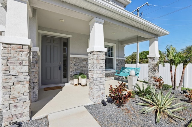 view of exterior entry featuring stone siding, stucco siding, a porch, and fence