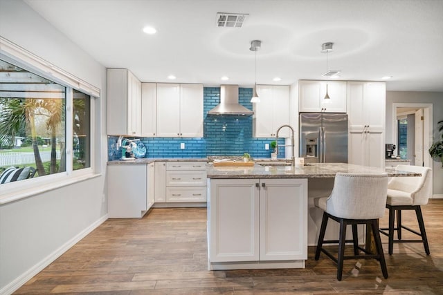 kitchen featuring stainless steel fridge with ice dispenser, visible vents, an island with sink, wood finished floors, and wall chimney exhaust hood