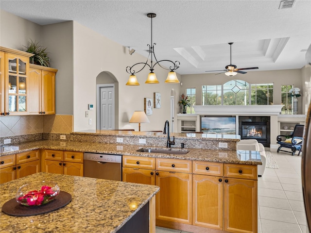 kitchen featuring light tile patterned floors, a sink, stainless steel dishwasher, a tray ceiling, and a glass covered fireplace