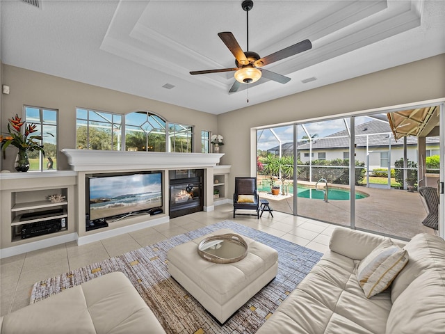 living room with a raised ceiling, a ceiling fan, a glass covered fireplace, a sunroom, and tile patterned floors