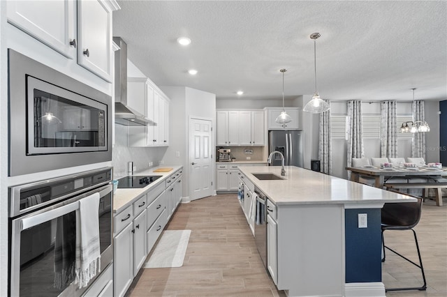 kitchen featuring light wood finished floors, white cabinets, wall chimney exhaust hood, stainless steel appliances, and a sink