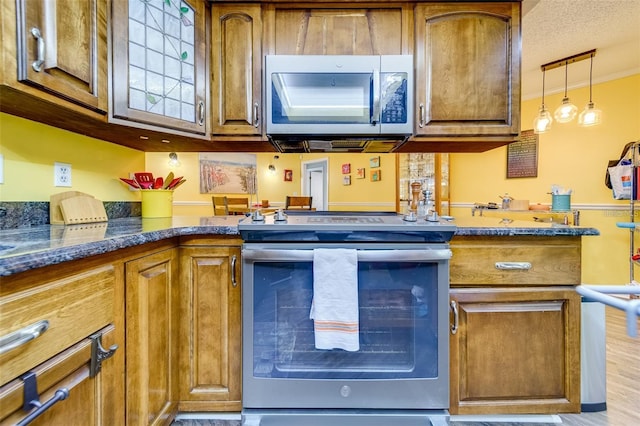 kitchen featuring hanging light fixtures, appliances with stainless steel finishes, brown cabinetry, dark stone countertops, and wood finished floors