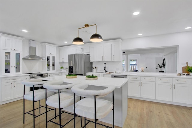 kitchen featuring a breakfast bar area, light wood-style floors, white cabinets, wall chimney range hood, and appliances with stainless steel finishes