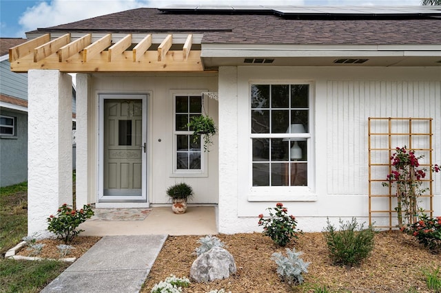 entrance to property with a shingled roof, solar panels, and stucco siding