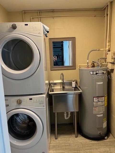 laundry area with electric water heater, laundry area, stacked washer / dryer, a sink, and concrete block wall