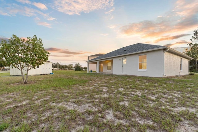 back of property at dusk featuring roof with shingles, an outbuilding, a lawn, and stucco siding