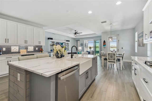 kitchen featuring backsplash, a center island with sink, a tray ceiling, light wood-style floors, and stainless steel dishwasher