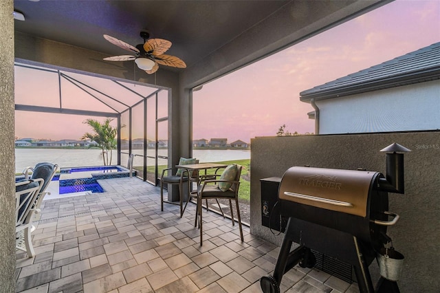 patio terrace at dusk featuring glass enclosure, a pool with connected hot tub, ceiling fan, a water view, and grilling area