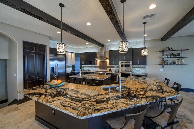 kitchen featuring stone tile floors, a sink, a large island, appliances with stainless steel finishes, and wall chimney range hood