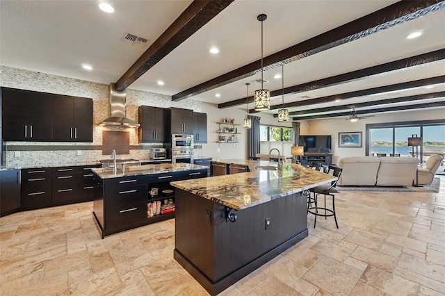 kitchen with wall chimney exhaust hood, a large island with sink, stone tile flooring, and beamed ceiling