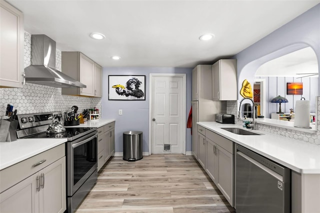 kitchen with light wood-type flooring, gray cabinetry, a sink, appliances with stainless steel finishes, and wall chimney exhaust hood
