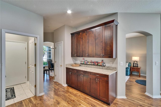 kitchen with a textured ceiling, arched walkways, light wood-style floors, dark brown cabinetry, and light stone countertops