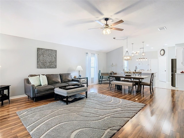 living room featuring baseboards, visible vents, wood finished floors, vaulted ceiling, and ceiling fan with notable chandelier