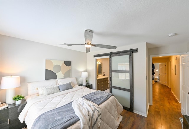 bedroom featuring a barn door, dark wood-style floors, baseboards, ceiling fan, and ensuite bathroom