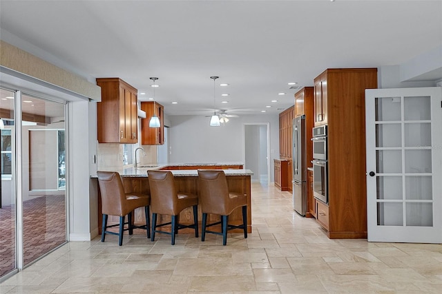 kitchen featuring brown cabinetry, a peninsula, a sink, stainless steel appliances, and stone finish floor