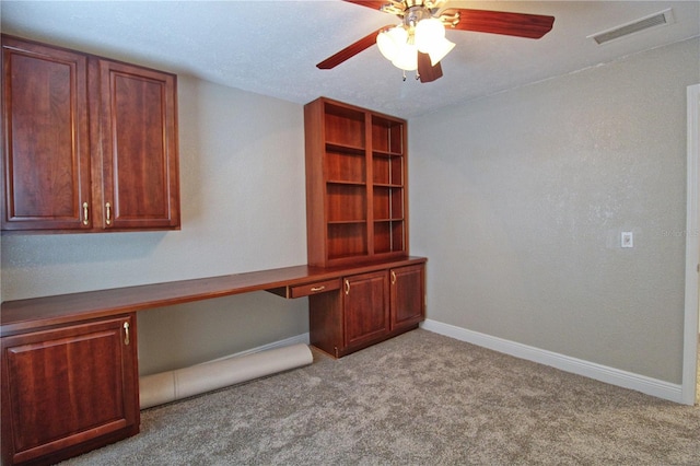 unfurnished office featuring built in desk, visible vents, a ceiling fan, carpet flooring, and baseboards